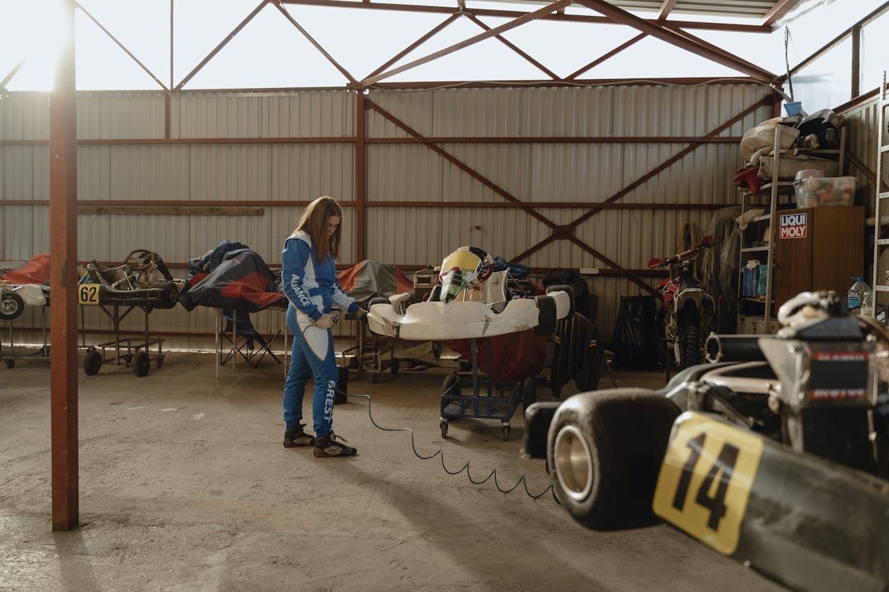 Female racer in blue racing suit prepares go-kart inside a sunlit garage, ready for the track.